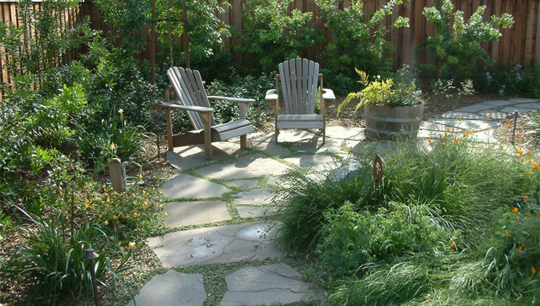Two weathered Adirondack chairs on a flagstone patio, groundcover growing between the stones, with a half-barrel planter and grasses alongside