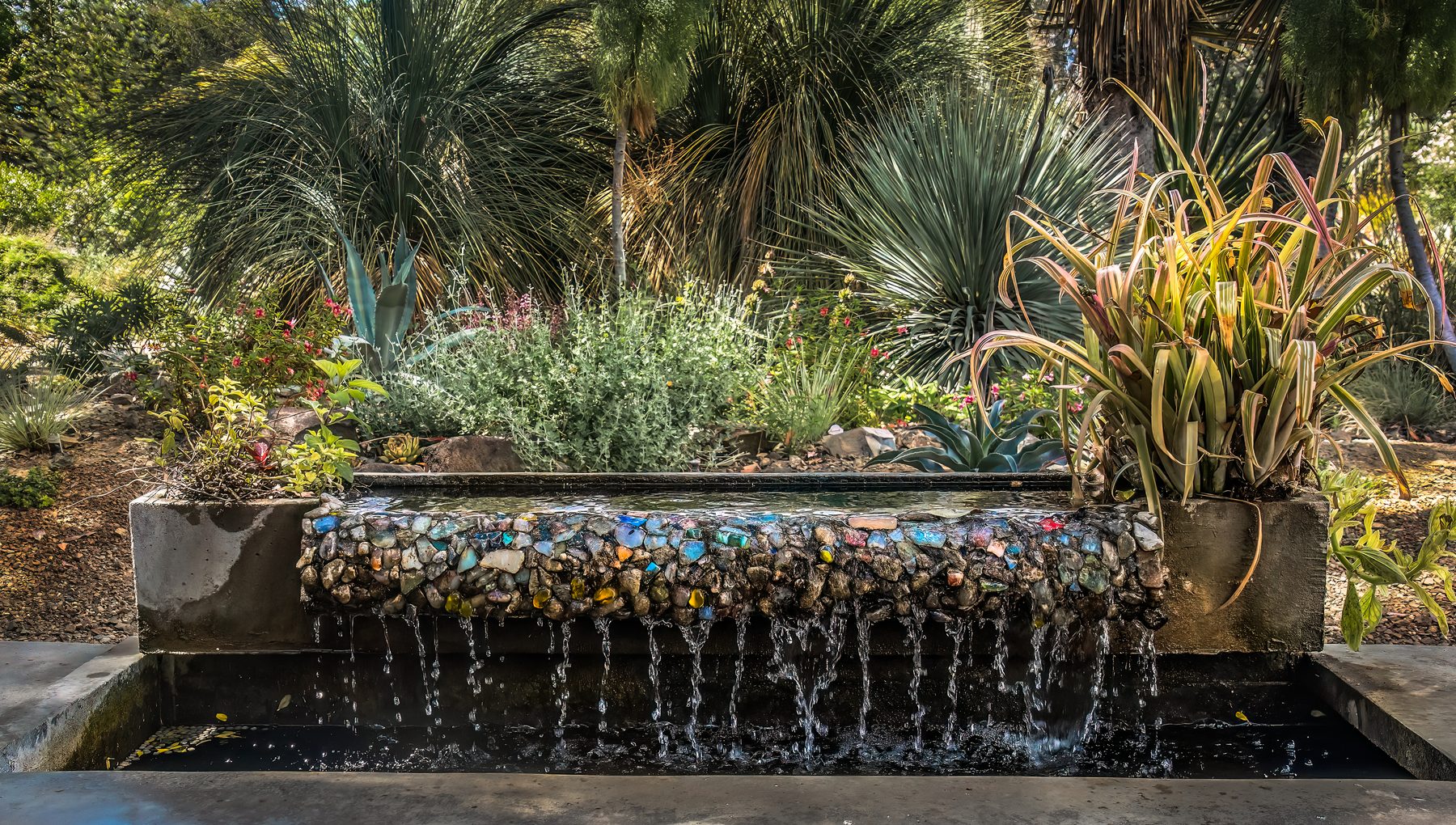 Water sheeting from a mosaic-faced wall into a long reflecting trough, with agaves and variegated flax planted above
